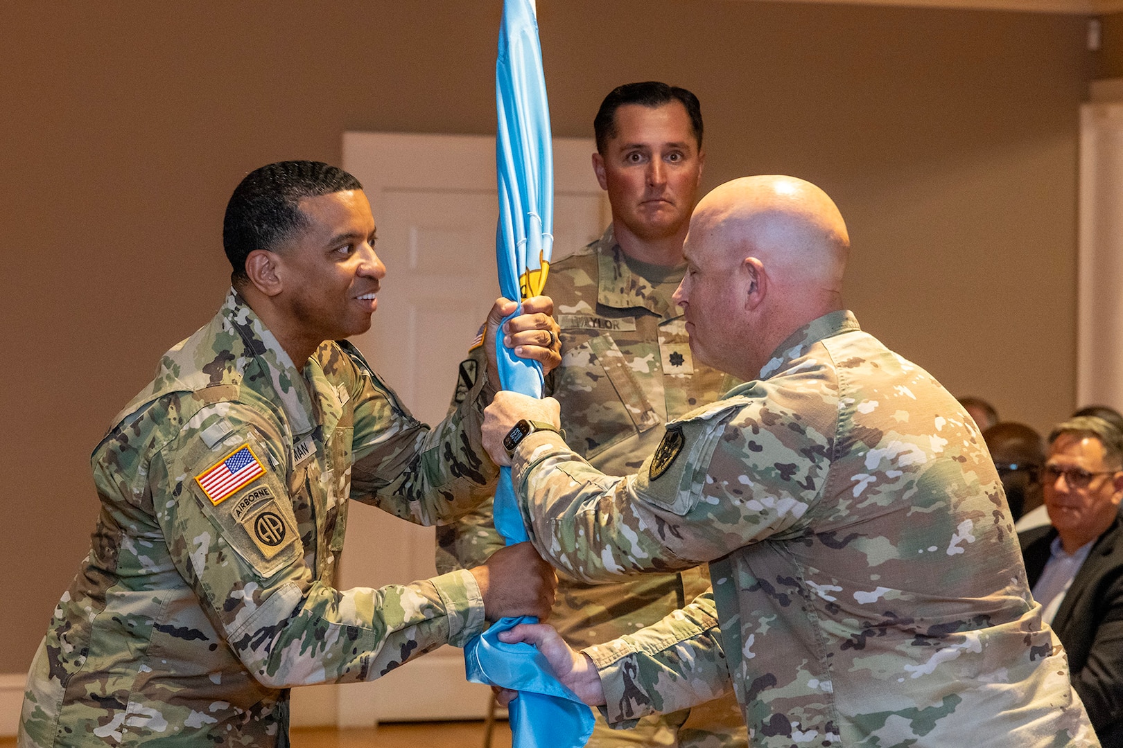 Two men wearing camouflage military uniforms stand on stage passing a guidon.