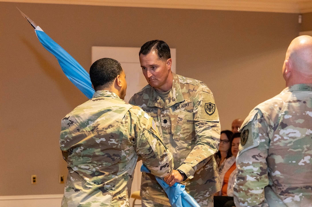 Two men wearing camouflage military uniforms stand on stage passing a guidon.