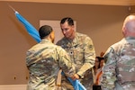 Two men wearing camouflage military uniforms stand on stage passing a guidon.