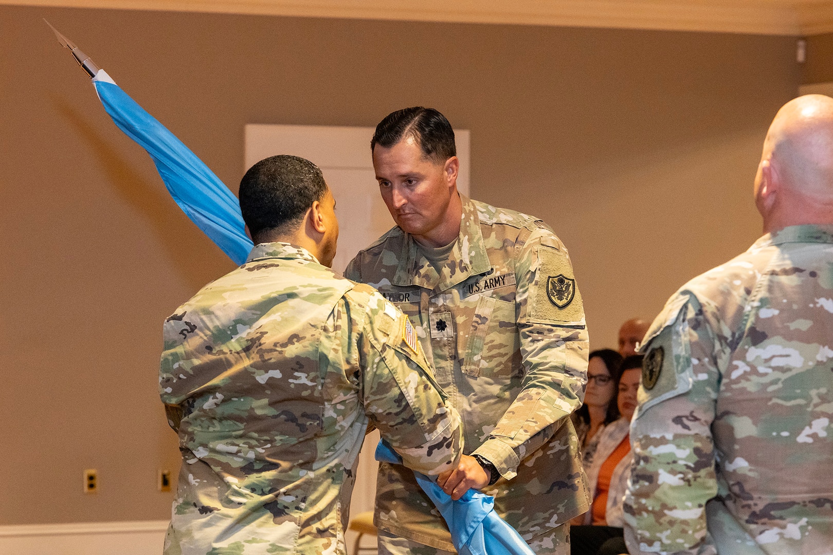 Two men wearing camouflage military uniforms stand on stage passing a guidon.