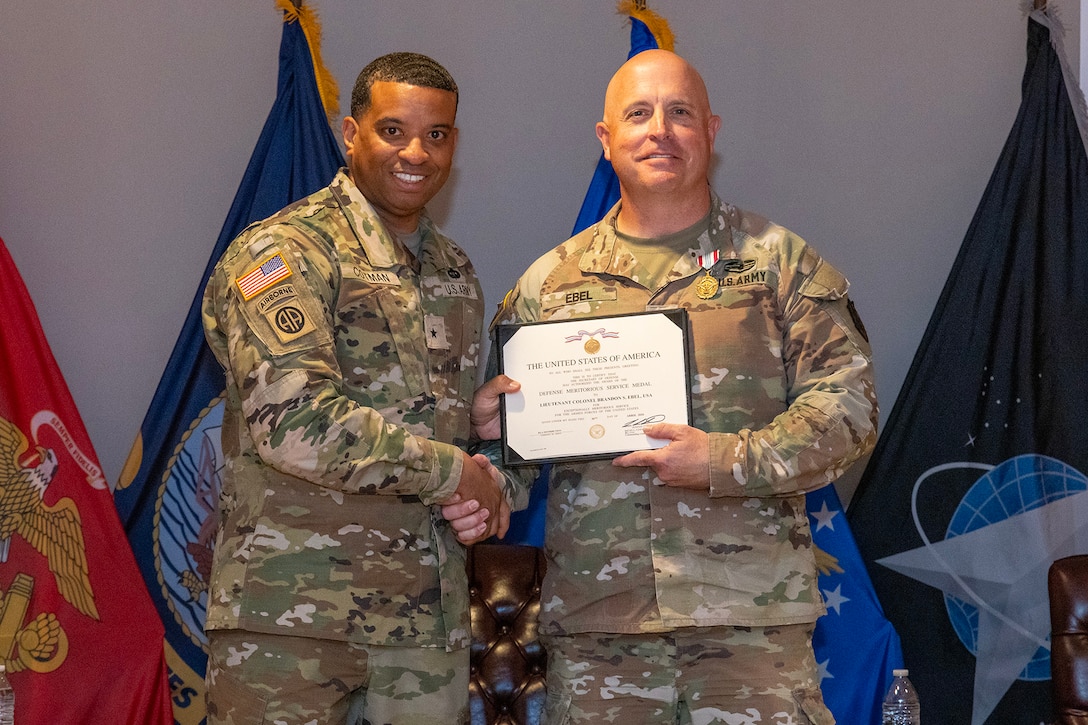 Two men wearing camouflage military uniforms stand on stage holding a certificate.