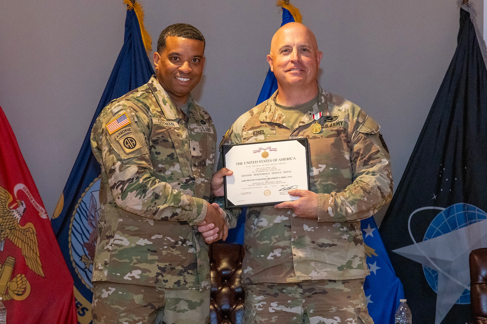 Two men wearing camouflage military uniforms stand on stage holding a certificate.