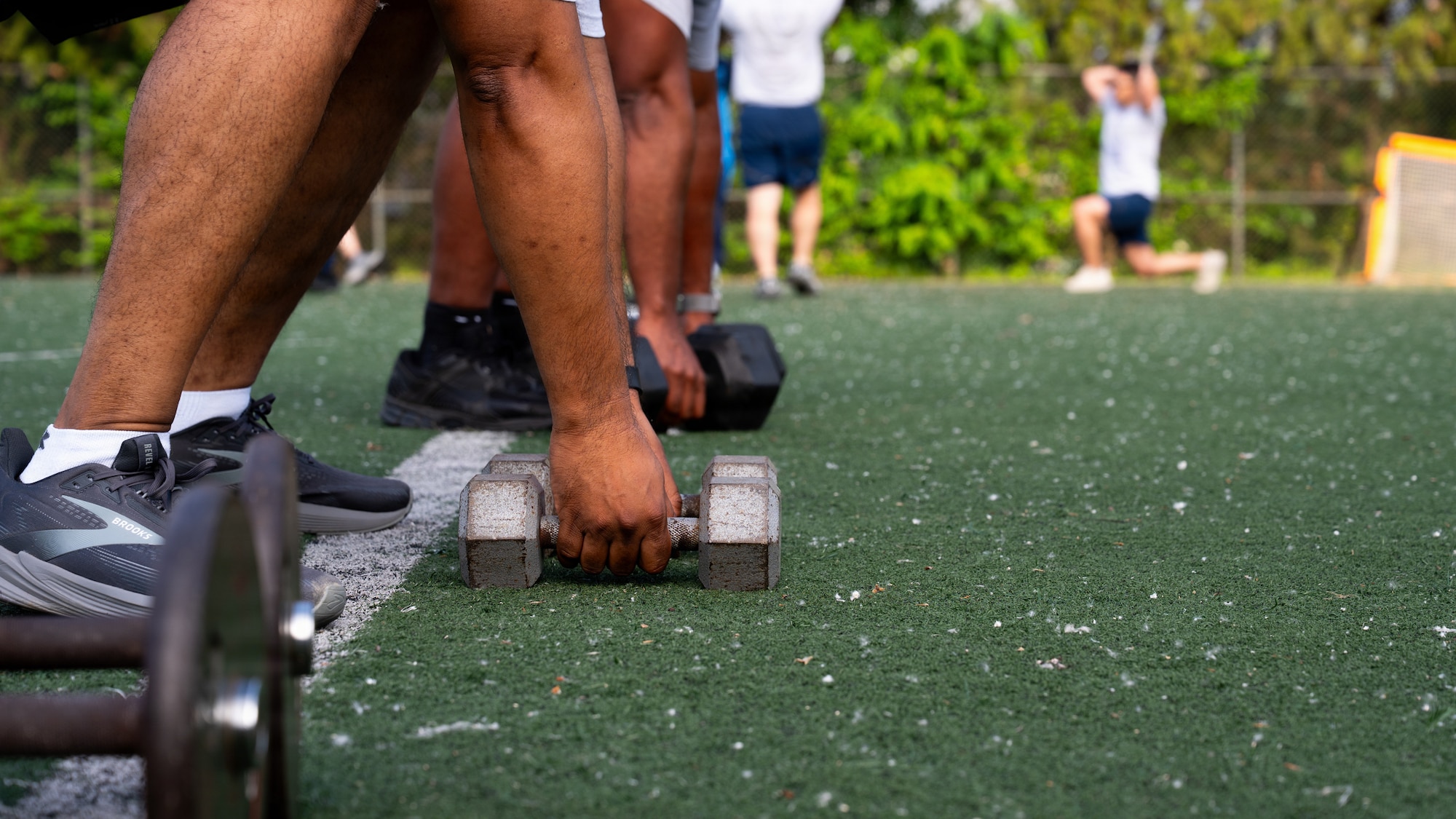 Hands reach down to grab dumbbells off of green turf. There's people in the background doing exercises