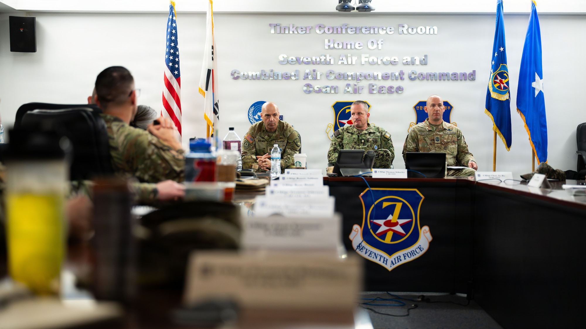 Three uniformed individuals are seated in the front of the a conference room in front of a U shaped table.
