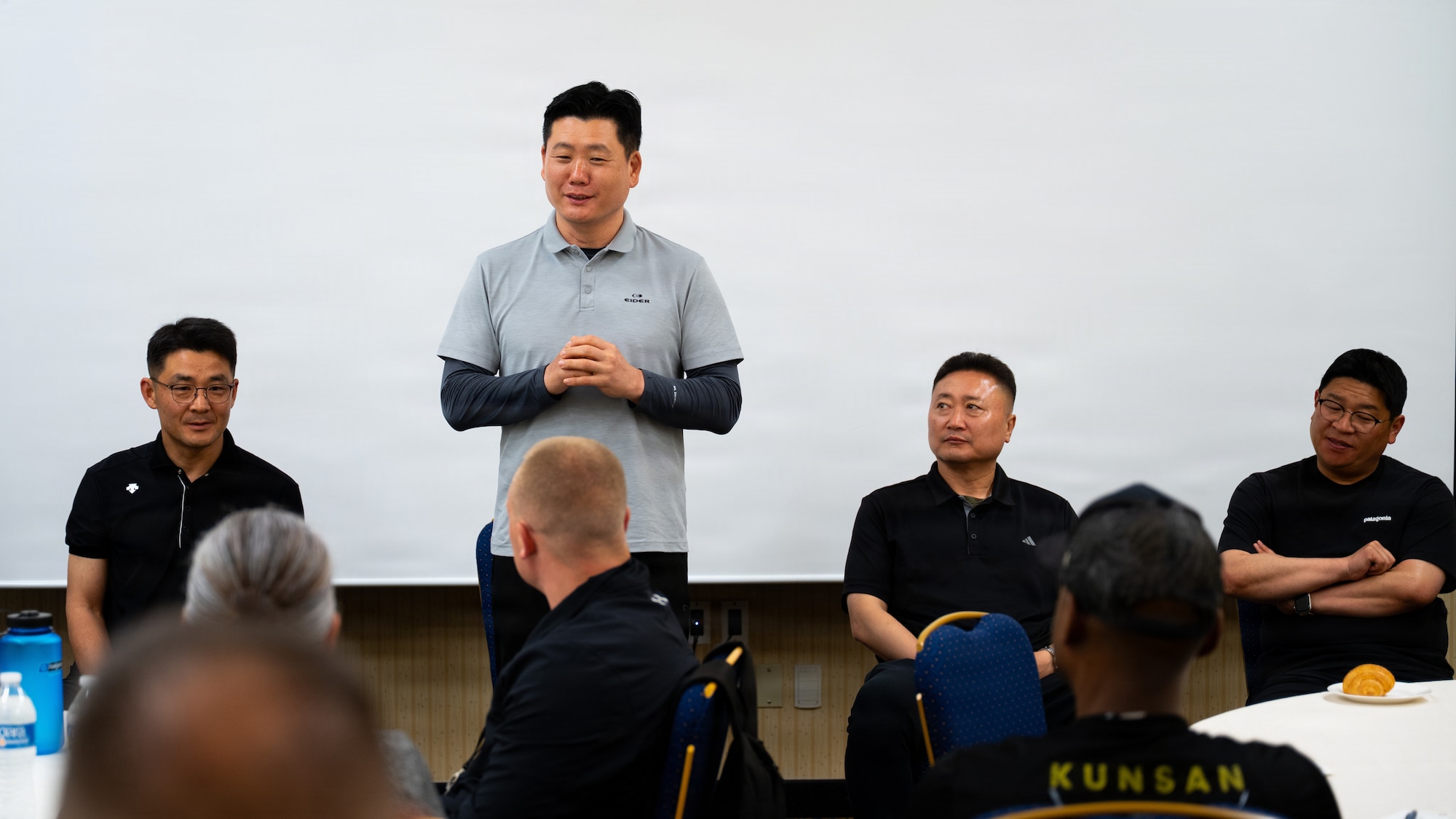 A group of four men, 3 sitting and 1 standing, are placed in front of a projector screen and talk to people in front of them.