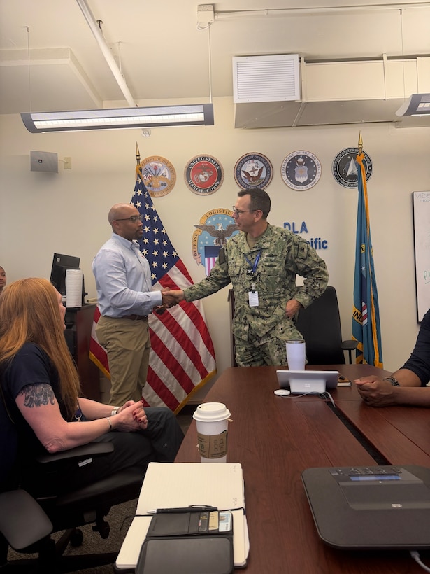 A civilian and Navy sailor shake hands at the front of a table