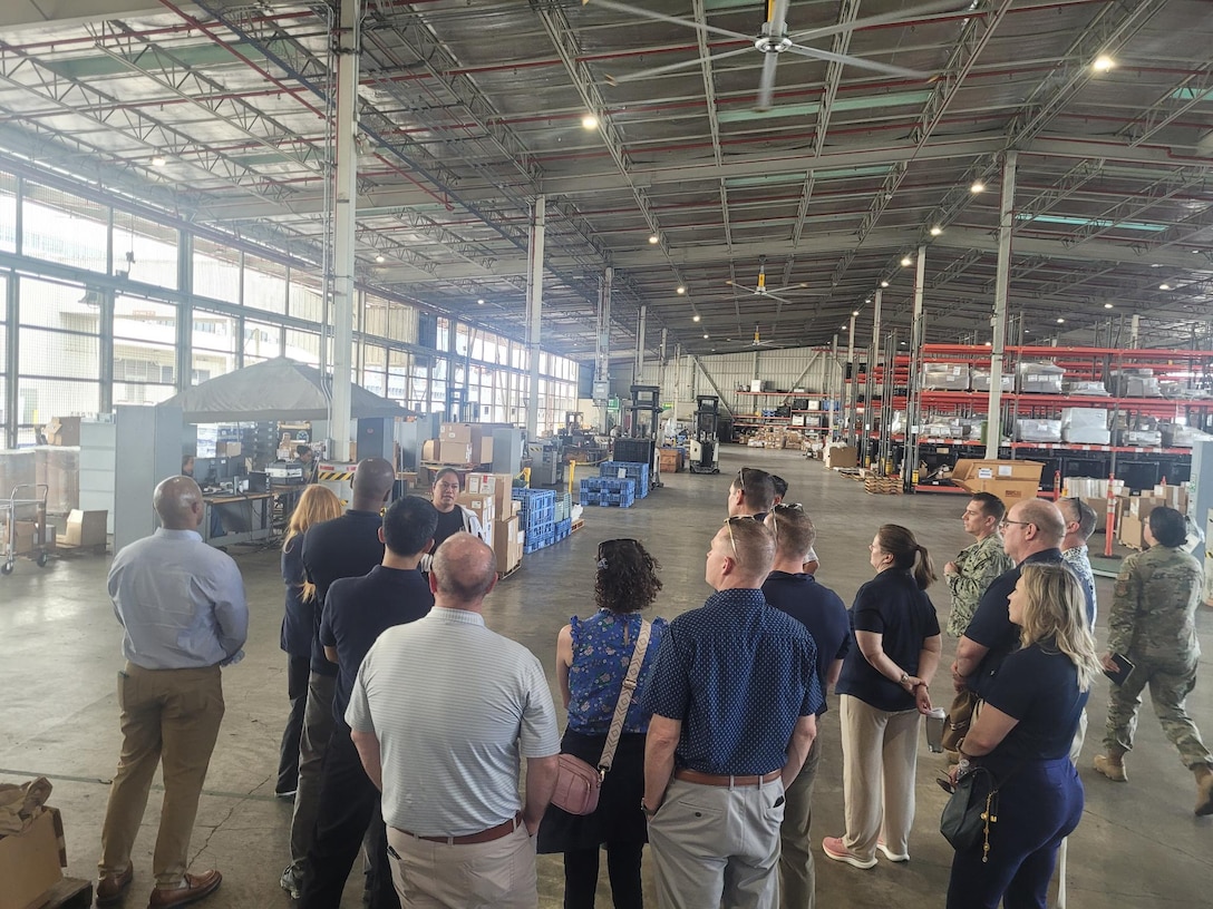 A group of people stand in a semi-circle with their backs toward the camera in a warehouse