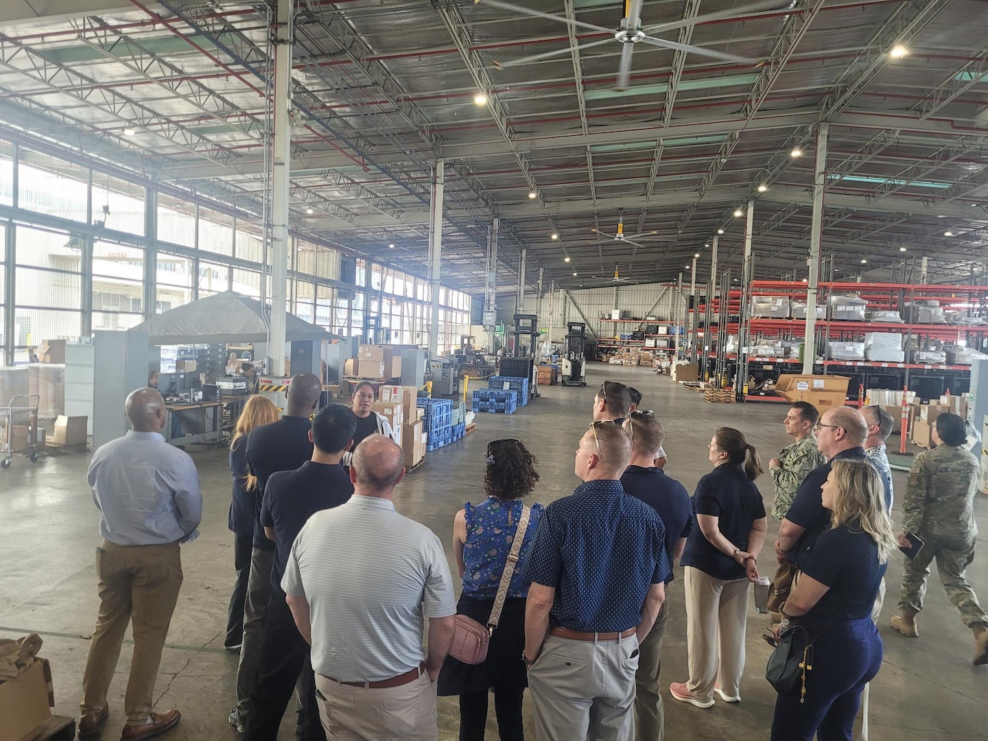 A group of people stand in a semi-circle with their backs toward the camera in a warehouse
