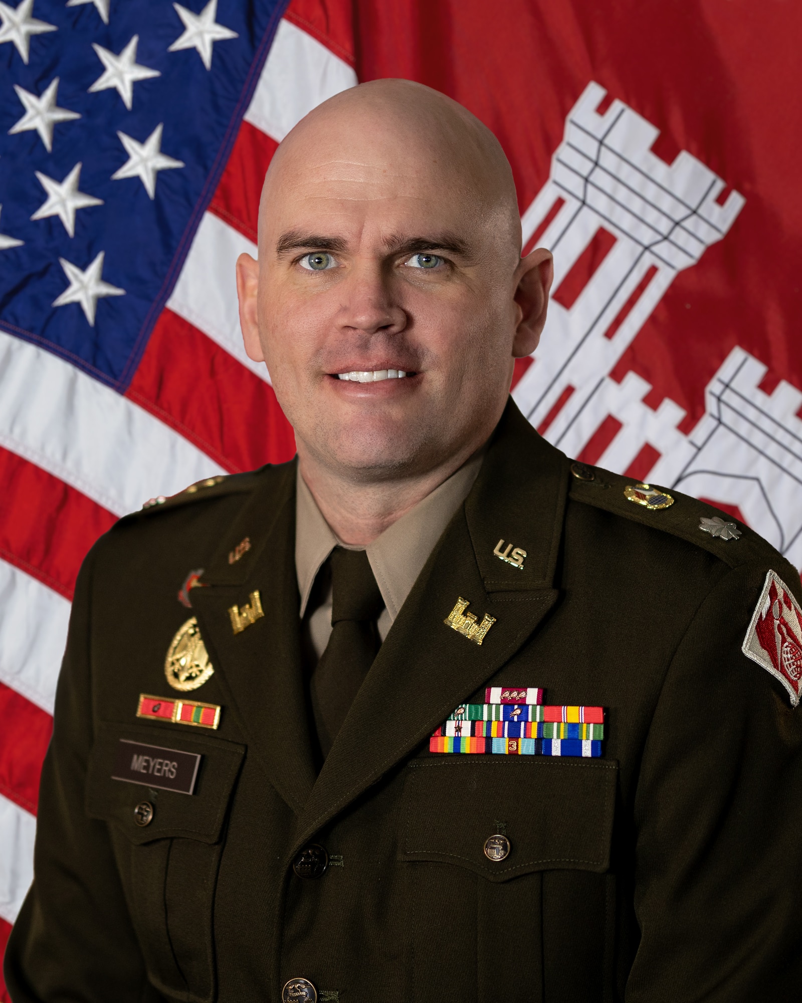 An image of a man wearing a dark green formal military uniform with an assortment of medals along his chest. Behind him is a red flag with a white castle, an American flag.