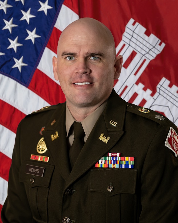 An image of a man wearing a dark green formal military uniform with an assortment of medals along his chest. Behind him is a red flag with a white castle, an American flag.