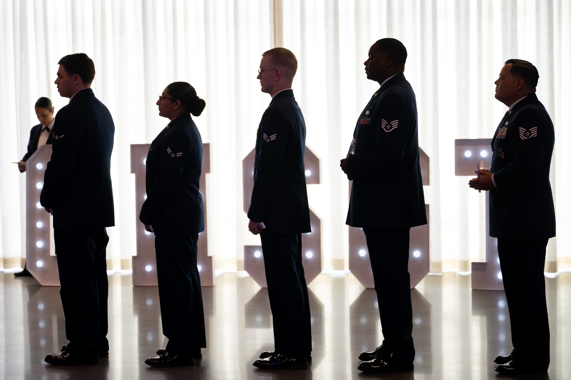 Five uniformed military personnel standing in a line against a bright curtain backdrop with illuminated letters behind them
