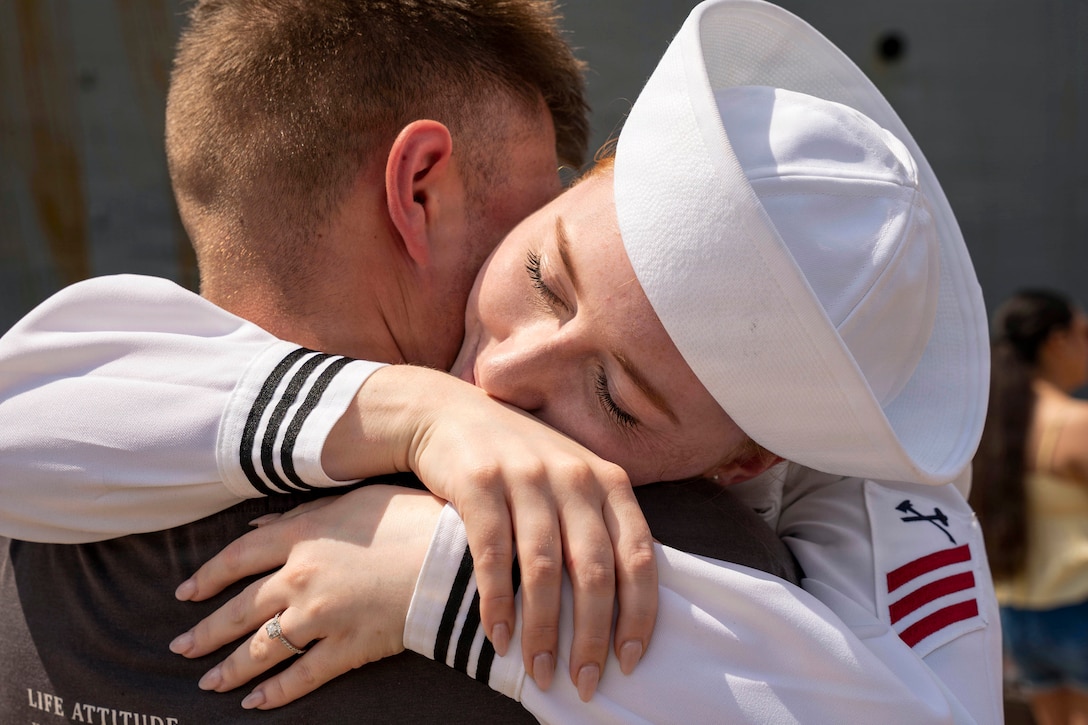 A sailor in a white uniform hugs a civilian outdoors by a moored ship.