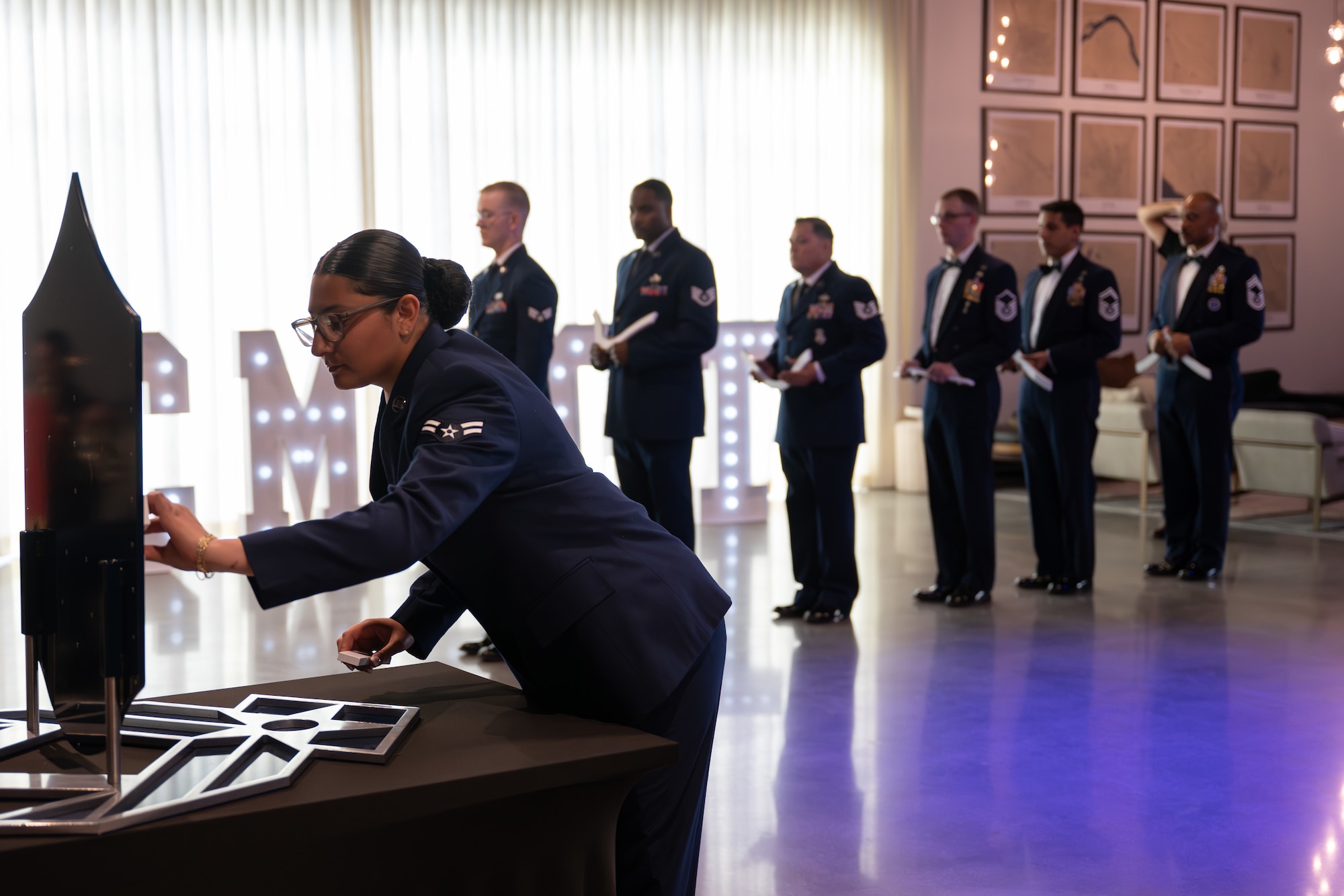 A group of uniformed military personnel standing in line with one female in uniform interacting with a display on a table in the foreground