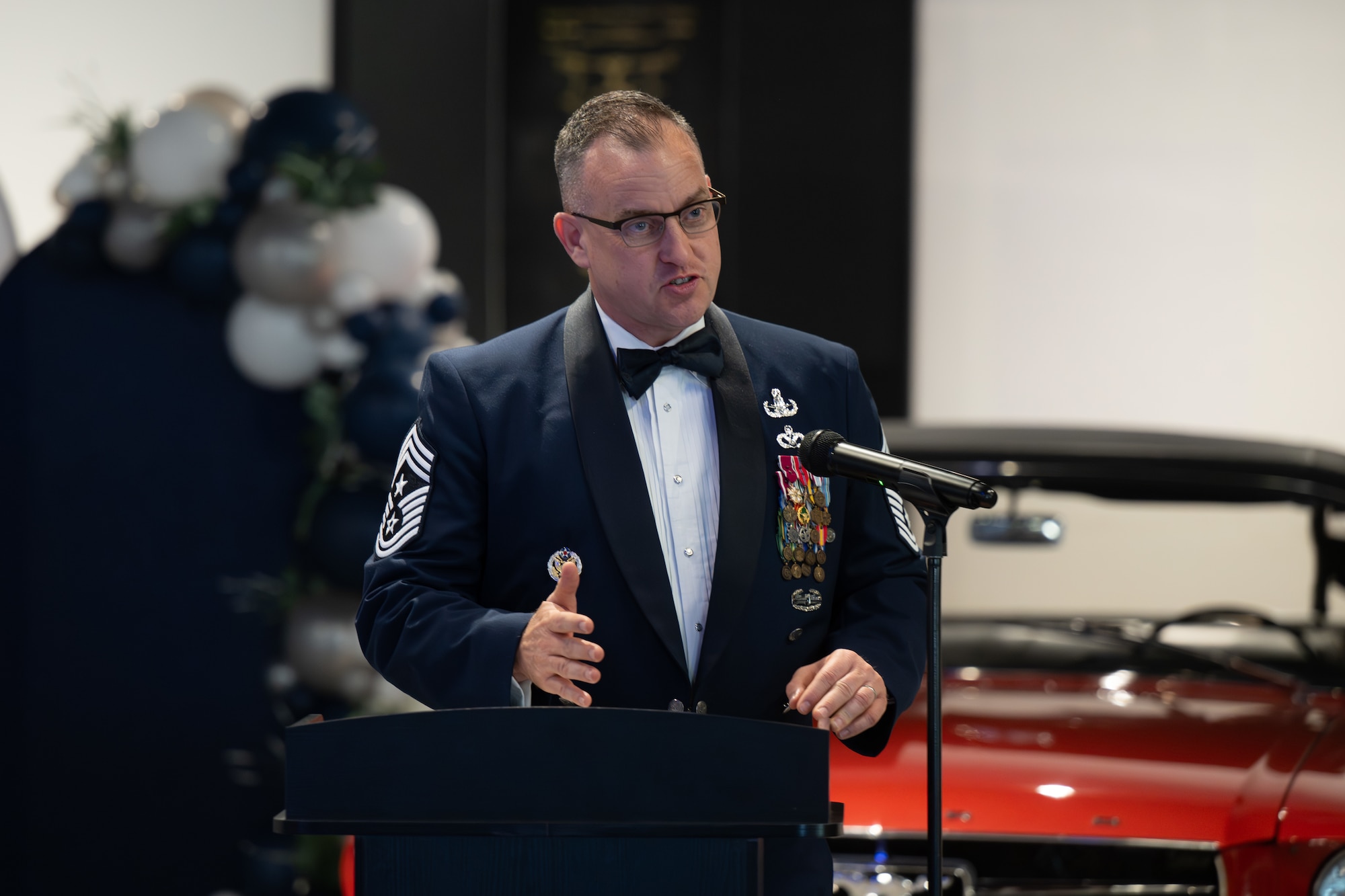 Man in formal military uniform speaking at a podium inside a showroom with a vintage red car