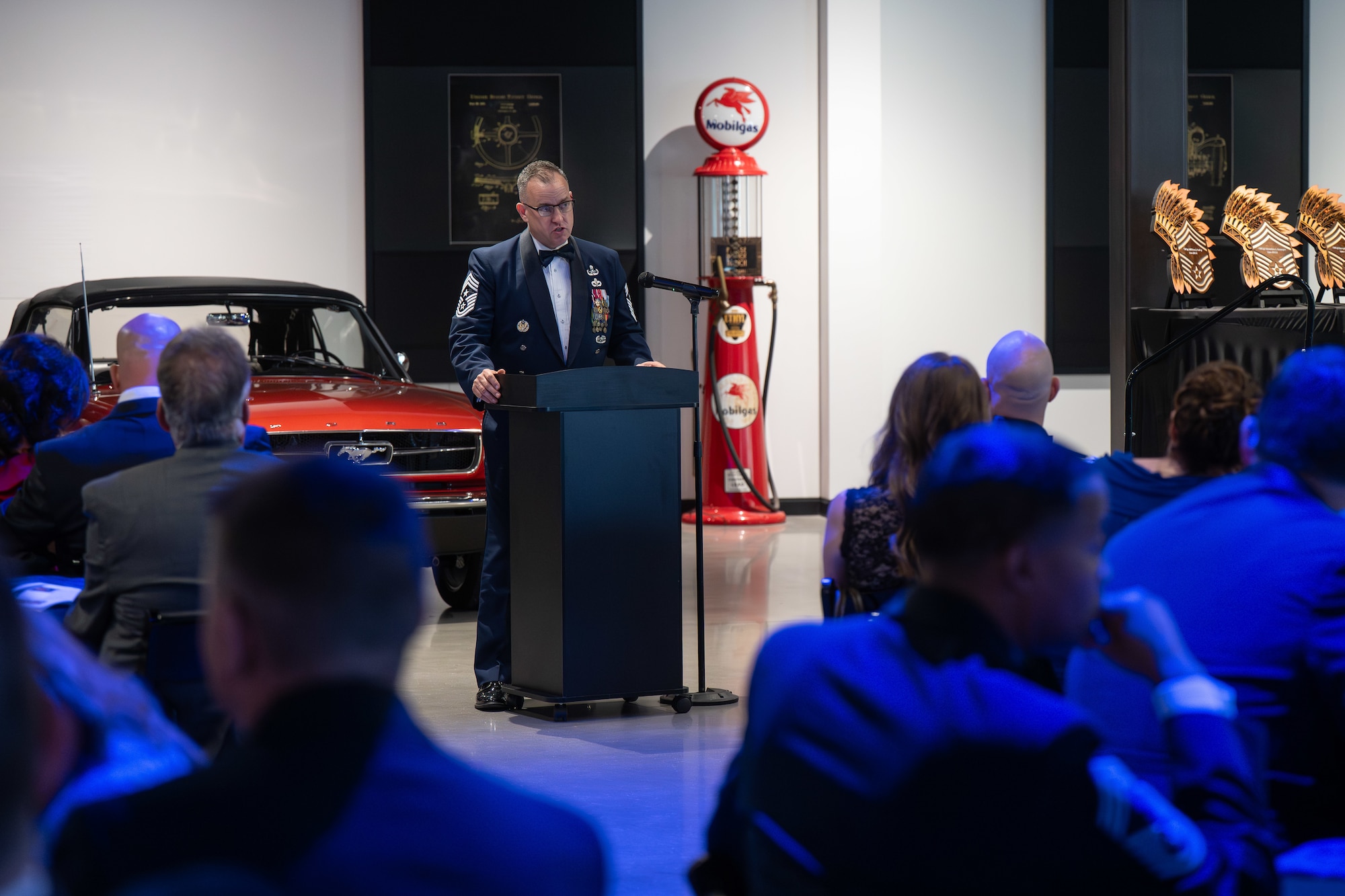 Man in formal military uniform speaking at a podium in front of an audience in side a showroom with a vintage red car and gas pump
