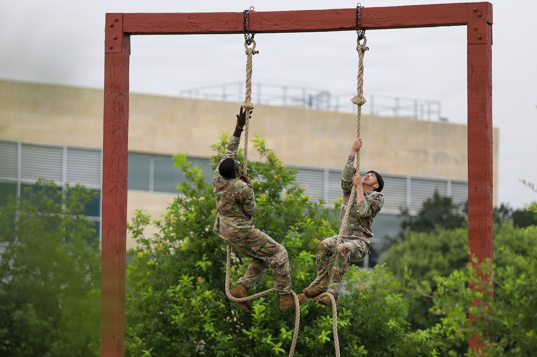 Two soldiers climb ropes hanging from a wood frame outdoors near a building.