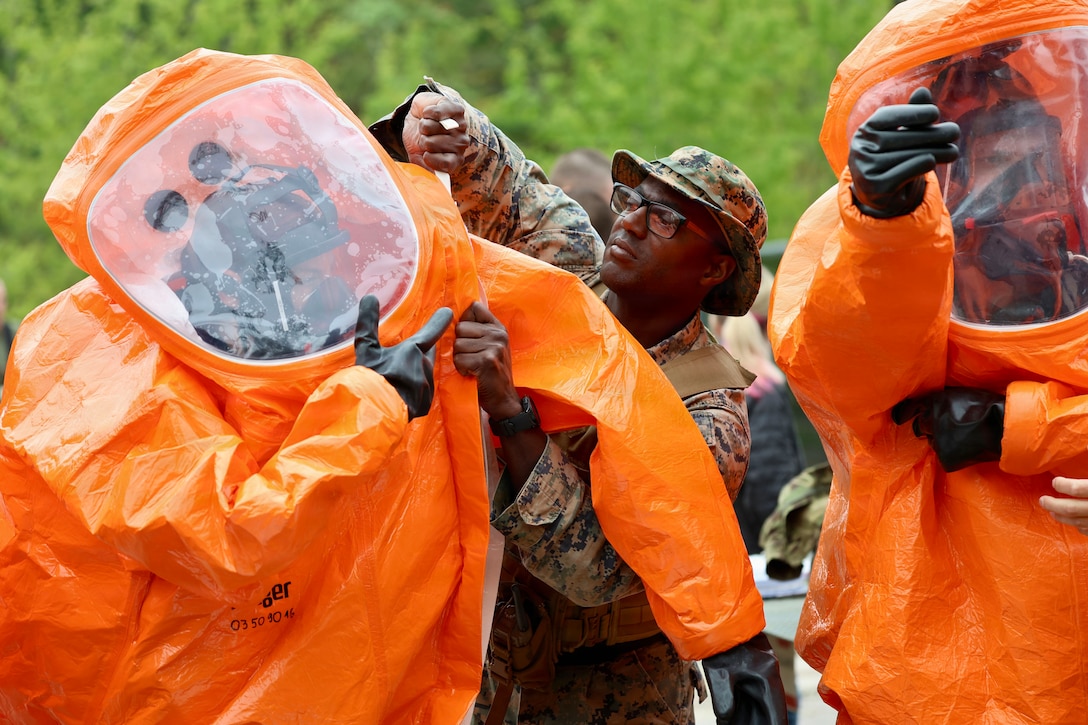 A Marine adjusts the sleeve of someone in an orange protective bodysuit while standing next to someone in identical gear outdoors.