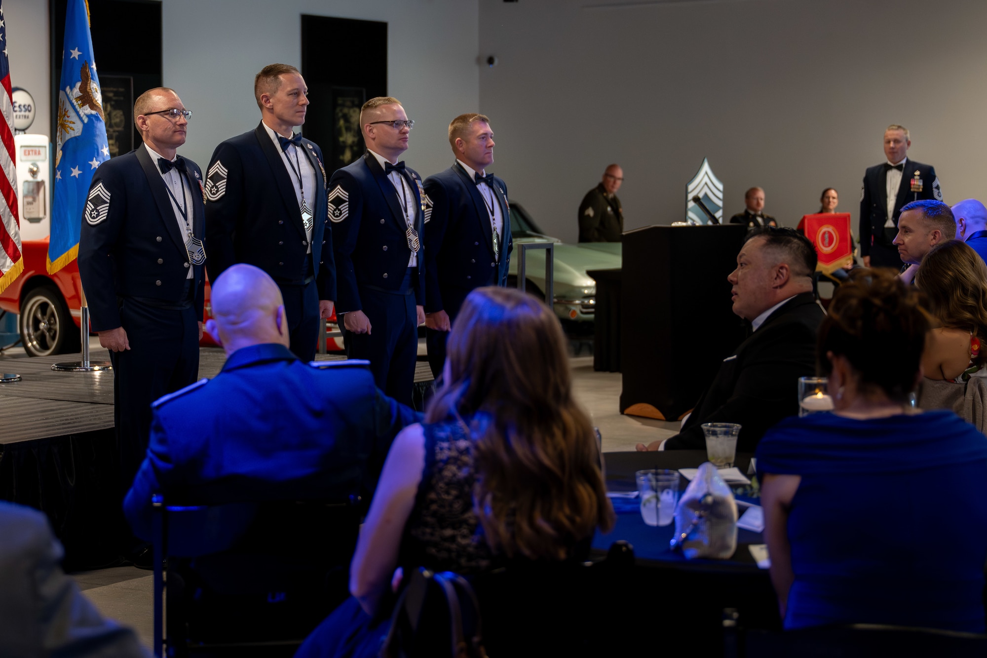 Four uniformed men stand in a row during a formal event in front of seated guests