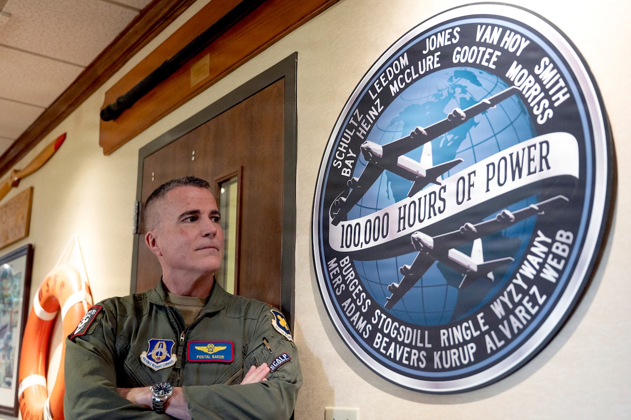A man wearing a flight suit stands in a hallway with his arms crossed looking at artwork on the wall of two military aircraft and a ribbon in the middle that reads, “100,000 Hours of Power.”