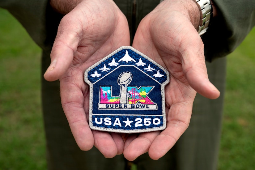 A pair of hands holds a patch with aircraft on the top that reads, “LIX Super Bowl, USA 250.”