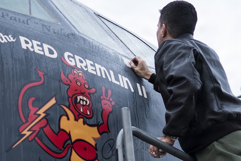 A man wearing a flight jacket peels paper off the side of a military aircraft while standing on a ladder.