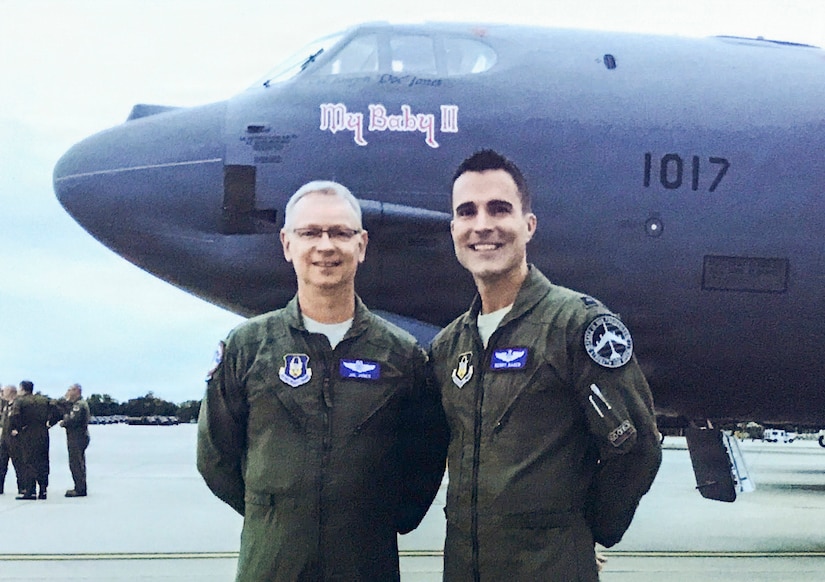 Two men wearing flight suits pose for a photo in front a military aircraft with writing on the side that reads,” My Baby II.”