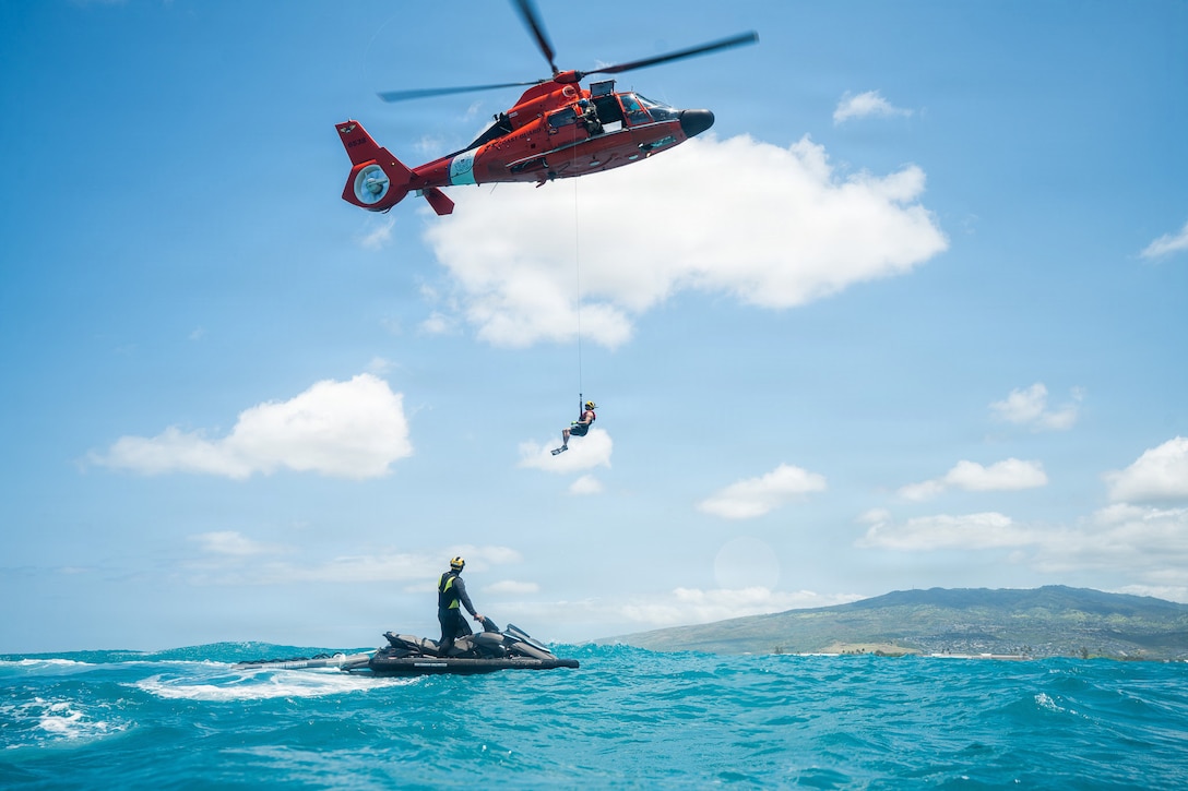 A Coast Guardsman on a personal watercraft in the water watches as a hovering orange helicopter hoists a swimmer attached to a line.