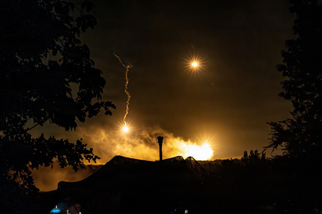 Flashes of light and smoke illuminate a dark sky above a howitzer in a field.