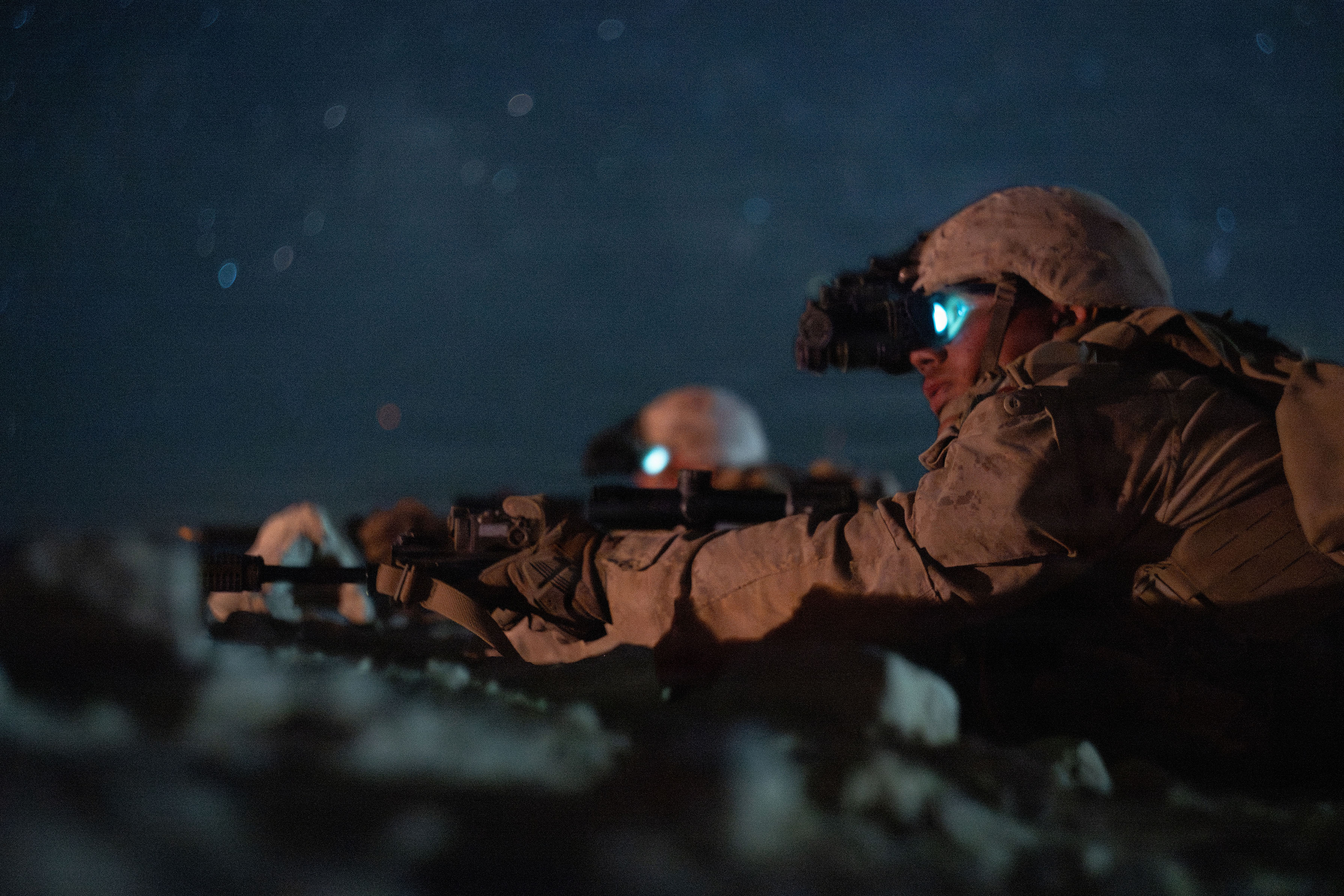 Marines wearing night-vision devices lie prone and point weapons under a dark starry sky.