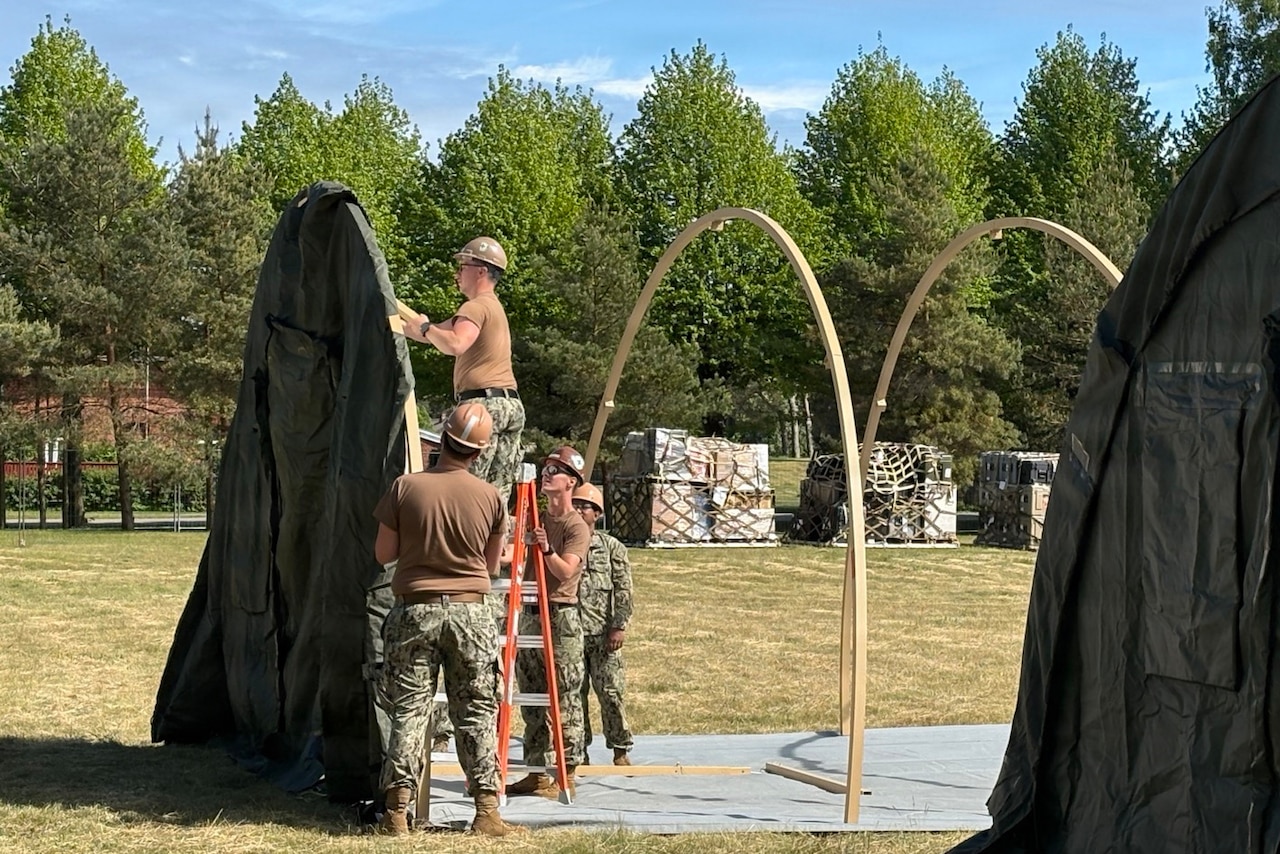 A man in a camouflage military uniform and hard hat stands on a ladder while putting up a large tent in a grassy area while several other people in similar attire hold the ladder and the tent frame.