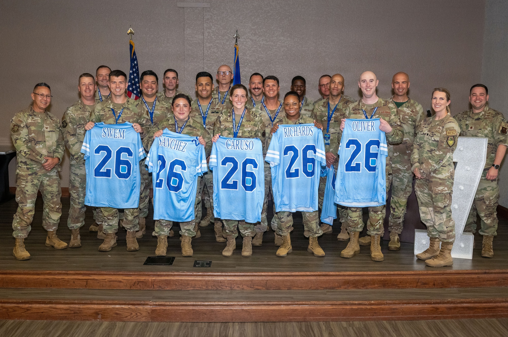 A group of people in military uniforms stand on a stage with five people holding up blue jerseys.