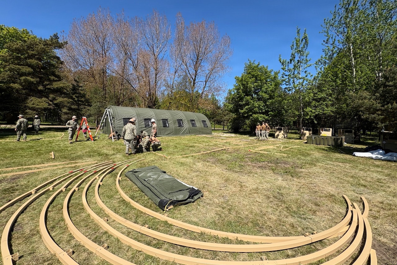 A dozen people in camouflage military uniforms construct large tents in a grassy area with trees in the background.; there are long pieces of curved wood lying on the ground in the foreground.