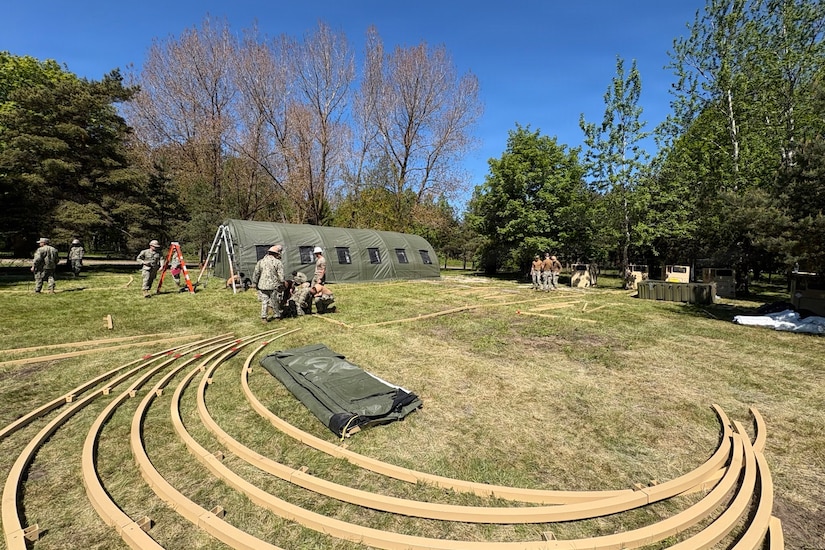 A dozen people in camouflage military uniforms construct large tents in a grassy area with trees in the background.; there are long pieces of curved wood lying on the ground in the foreground.
