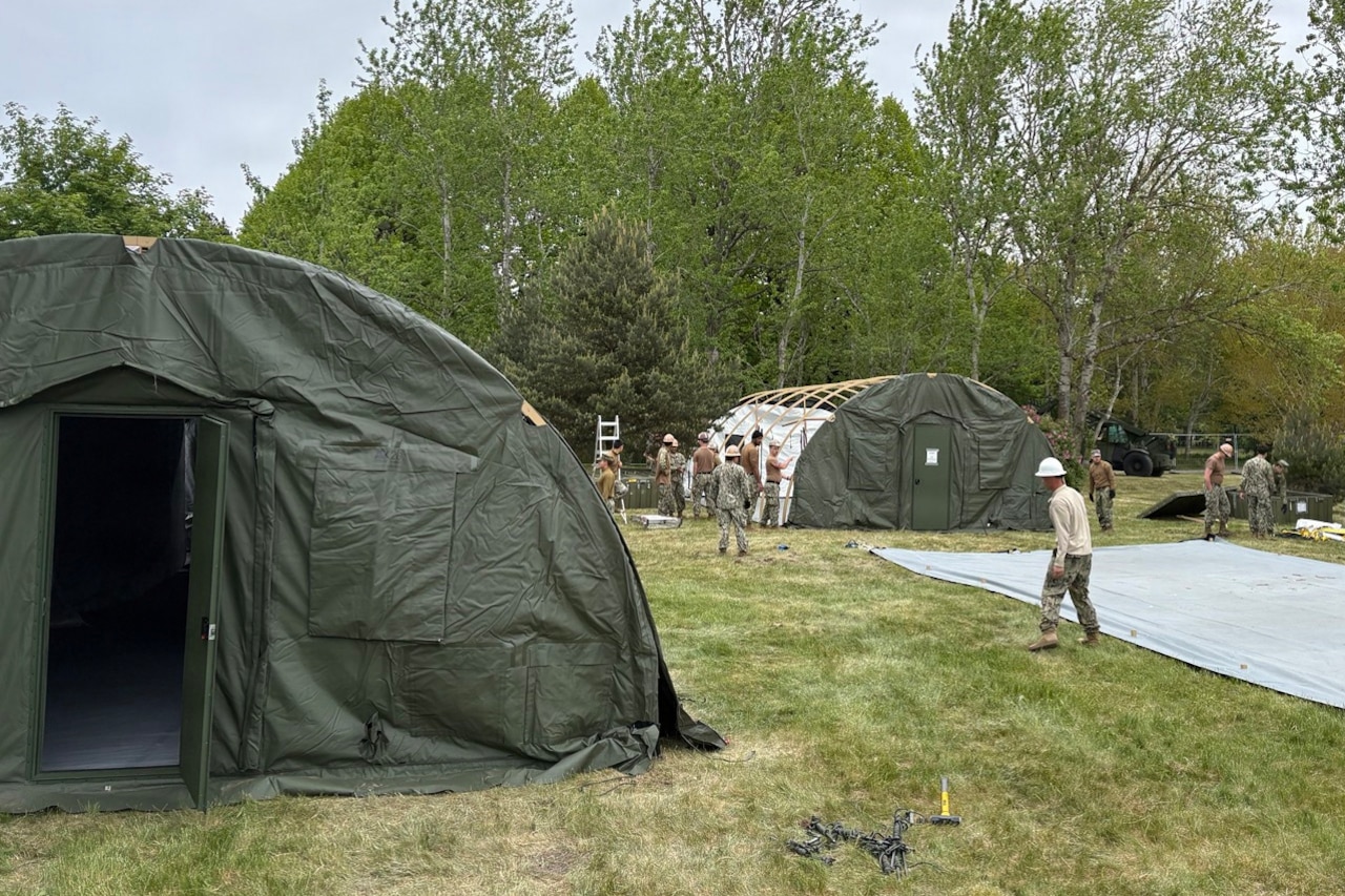 A dozen people in camouflage military uniforms construct large tents in a grassy area with large trees in the background.