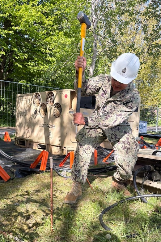 A man in a camouflaged military uniform and a white hard hat hammers a stake into the group in a grassy area as another man in similar attire observes; there is construction equipment in the background.
