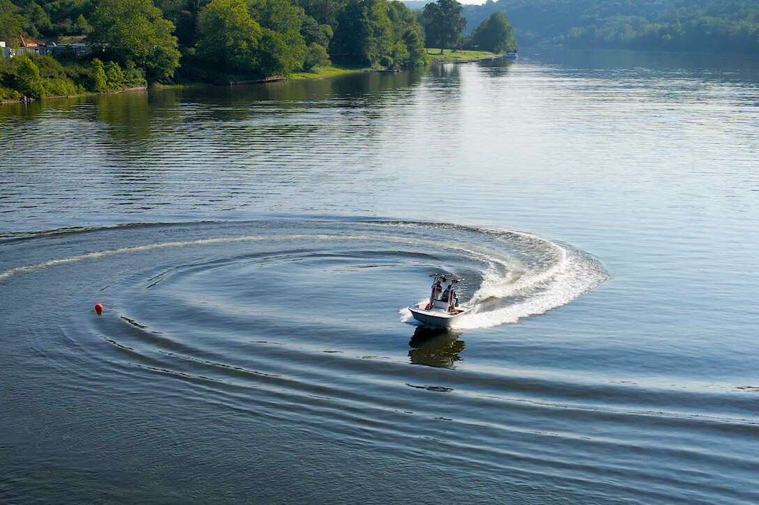 A white motorboat with people on board makes a sharp turn on a calm river, creating a large, swirling wake. A red buoy floats in the water nearby, and the distant shore is covered in green trees.