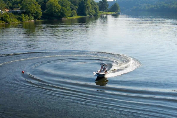 A white motorboat with people on board makes a sharp turn on a calm river, creating a large, swirling wake. A red buoy floats in the water nearby, and the distant shore is covered in green trees.