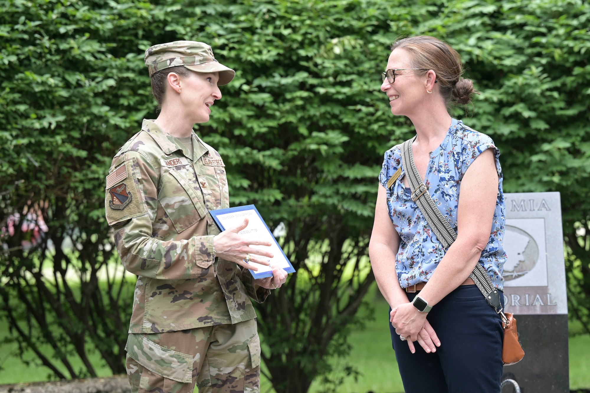 A woman in a camouflage uniform smiles as she presents a certificate to another woman wearing civilian attire