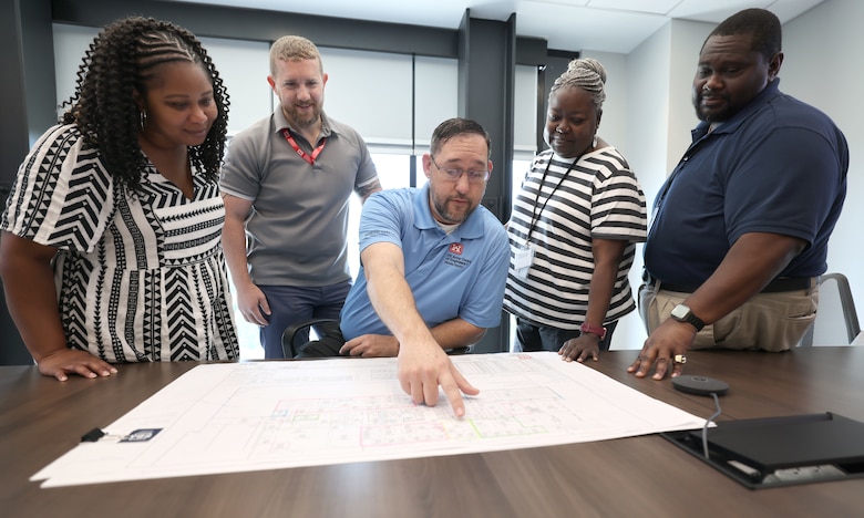 Picture of a man point at a blueprint with people looking on.