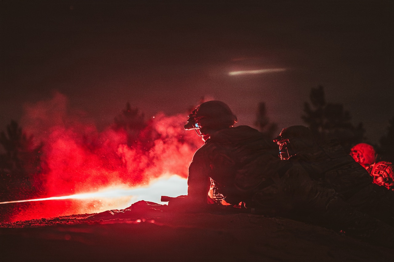 Soldiers lie prone and fire weapons, creating red light and smoke against a dark sky.