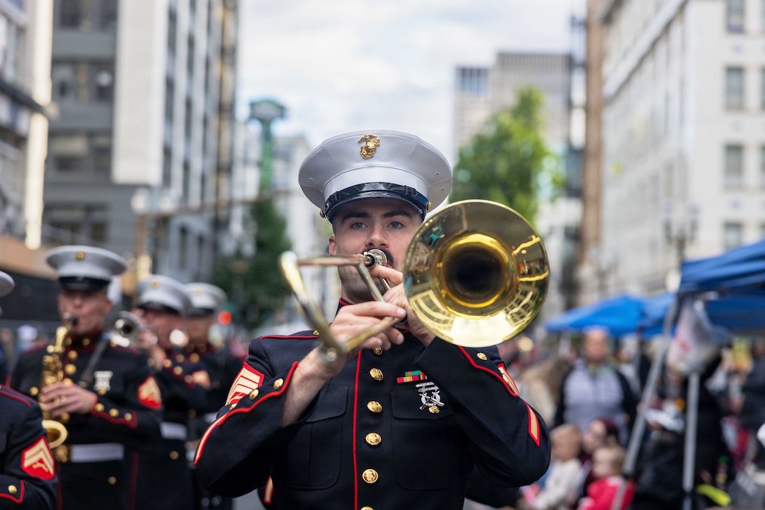 A close-up of a Marine in ceremonial dress playing a trumpet with band members during a parade with tall buildings in the background.