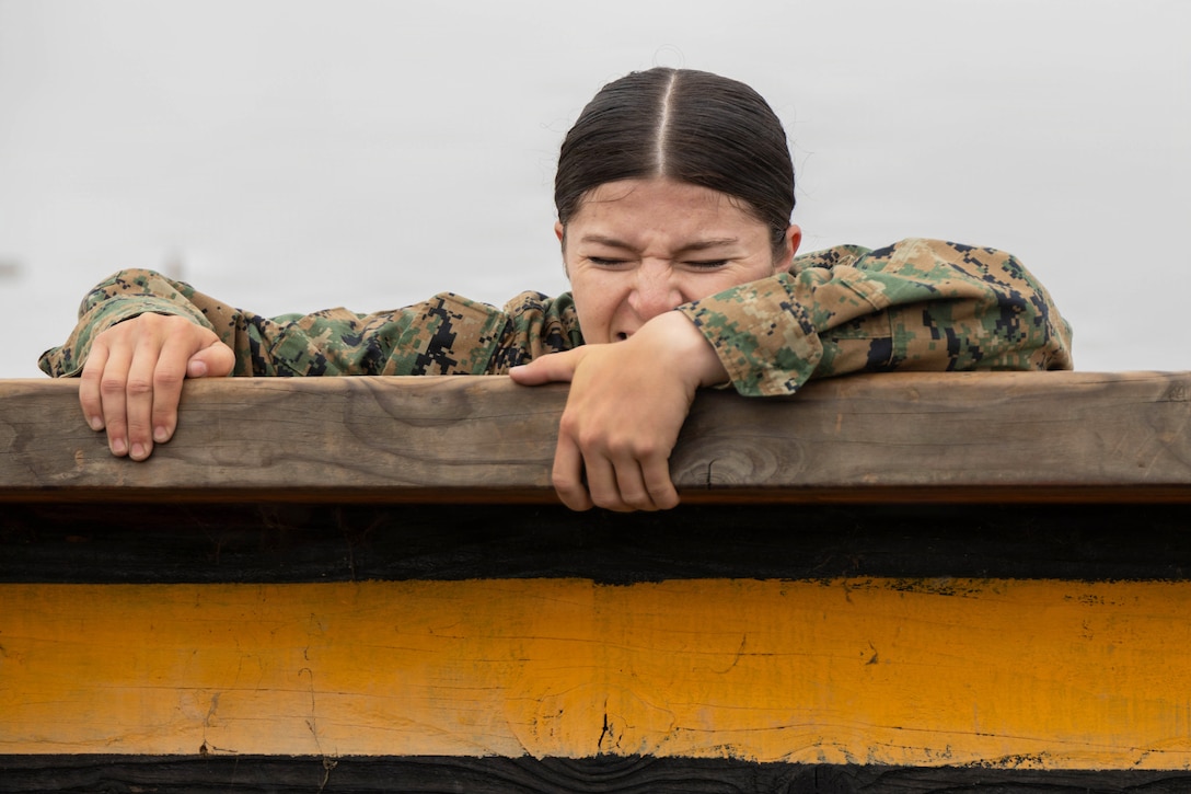 A close-up of a midshipman straining their face while climbing over an obstacle against a gray sky.