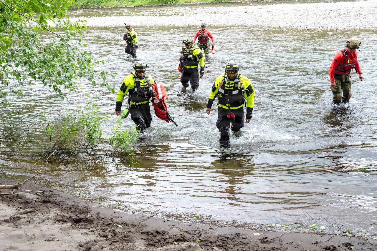 Air Force pararescuemen and first responders in protective gear walk through a river in a wooded area during the day.