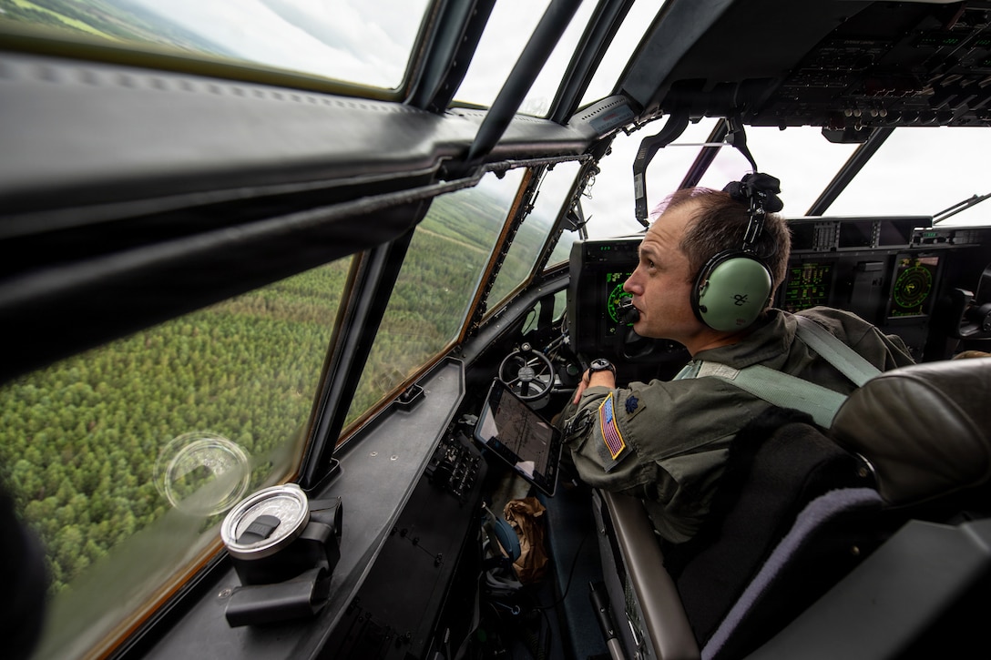 An airman in an airborne aircraft looks out of a cockpit at a field of trees.