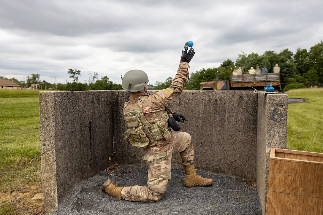 A soldier tosses a grenade from behind a concrete structure toward targets in an old pickup truck in a field.