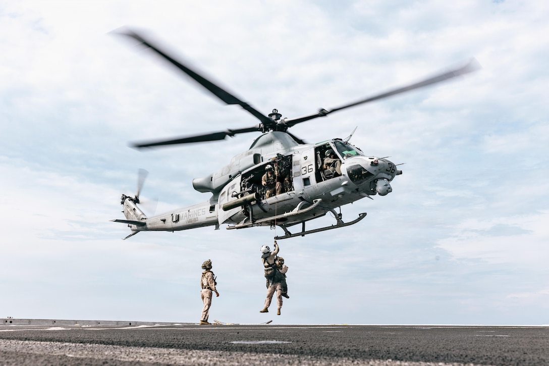 Two service members are hoisted on an airborne helicopter hovering over a ship at sea as a fellow service member watches.