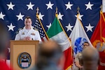 A man wearing a white dress U.S. Navy military uniform stands on stage indoors behind a podium speaking to a group of people with flags behind him.