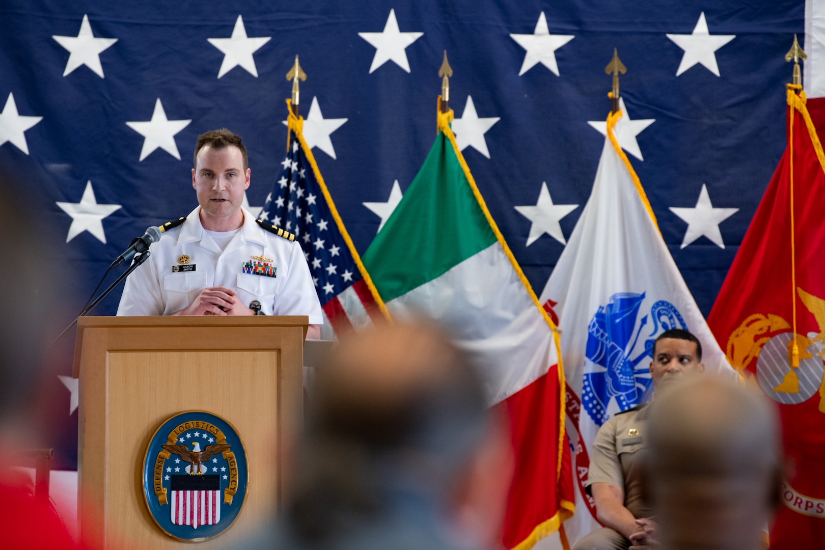 A man wearing a white dress U.S. Navy military uniform stands on stage indoors behind a podium speaking to a group of people with flags behind him.