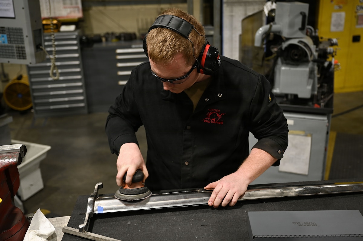 A man wearing a black shirt and protective eye gear uses a power sander to sand a large sword. Heavy machinery is behind him.