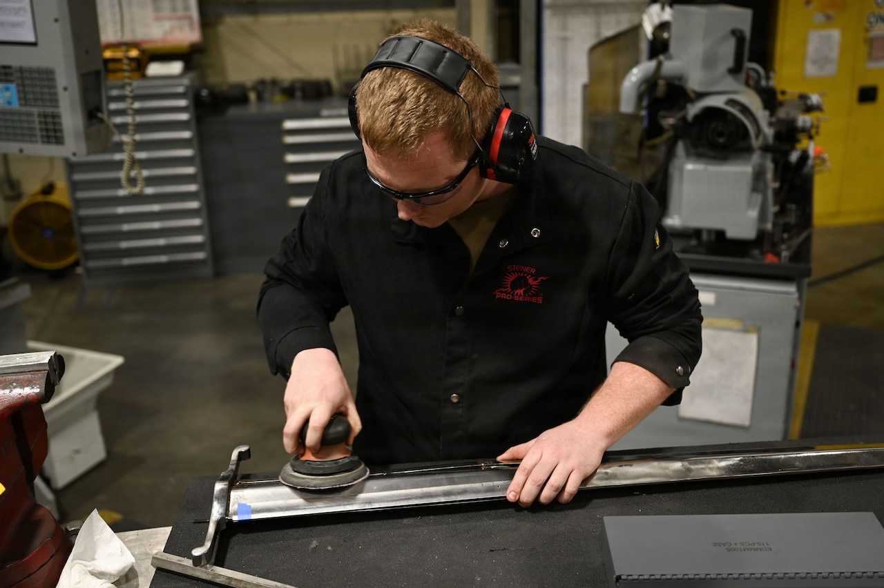 A man wearing a black shirt and protective eye gear uses a power sander to sand a large sword. Heavy machinery is behind him.